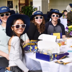 A group of smiling young adults sits at a table, each wearing white sunglasses 和 matching blue hats. They are surrounded by gift boxes, pamphlets, 和 floral arrangements. The setting appears to be a casual event or gathering.